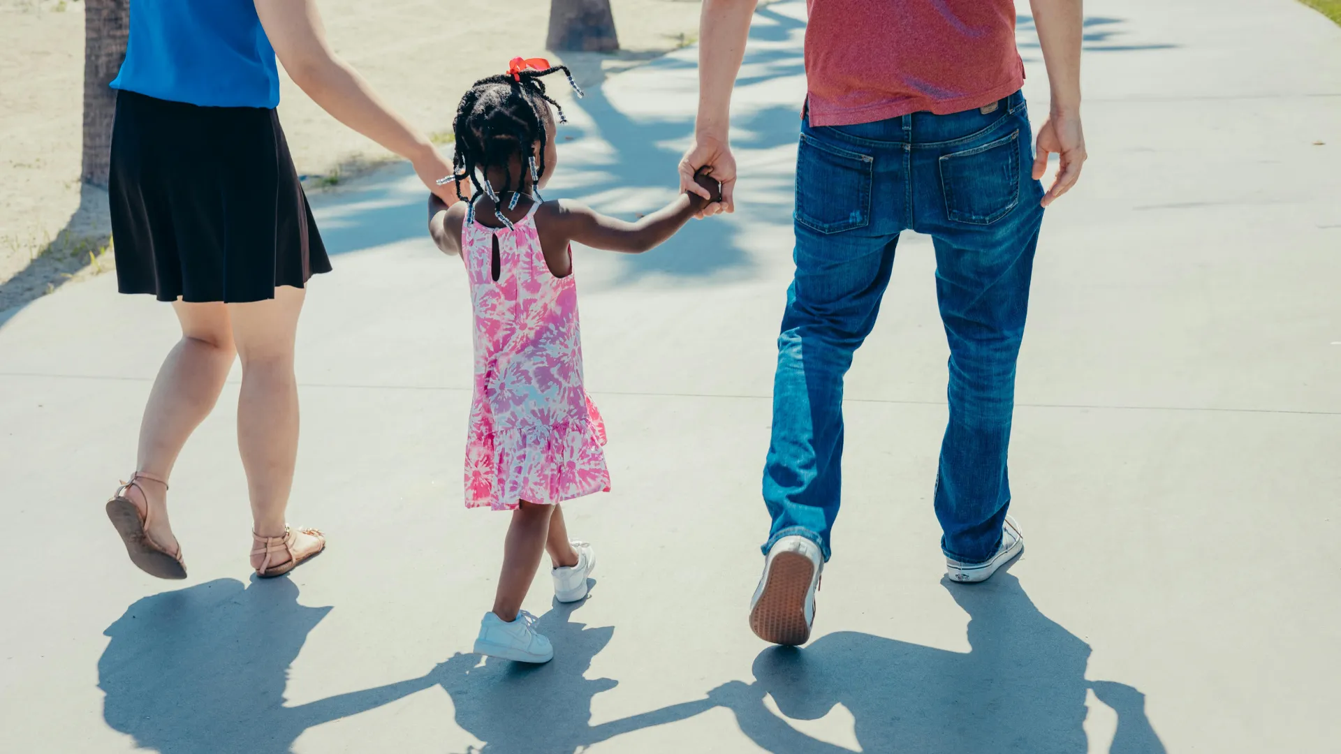 Multiracial family walking down the street with a little girl in the middle holding her parent's hands.