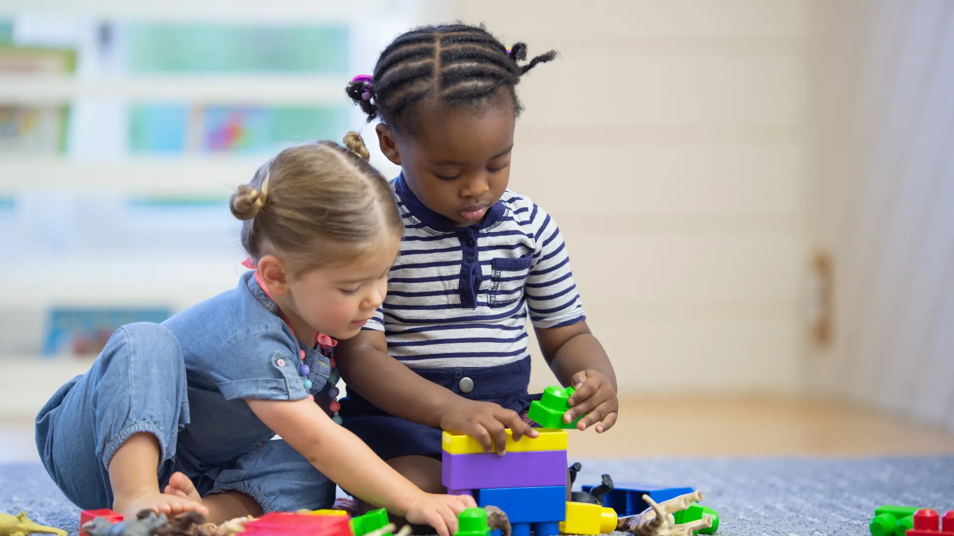 Two young children play together with colorful building blocks on a classroom floor.