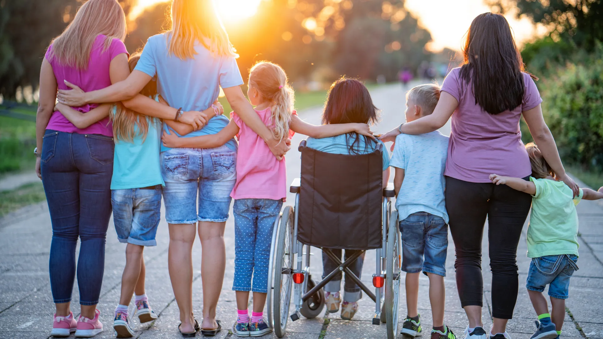 Inclusive foster family walking in the park Two adults and several children, including a child using a wheelchair, walk together along a park path at sunset.