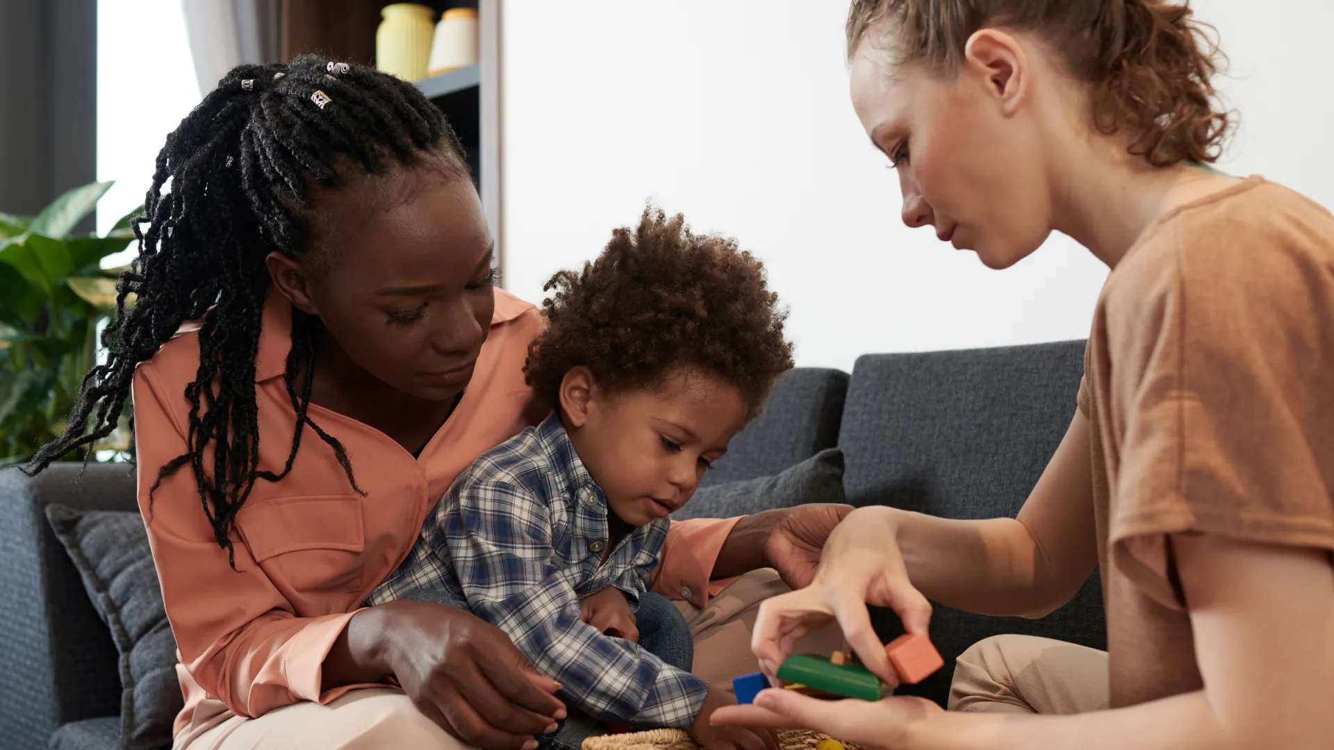 Foster Care- Supportive play with chill Two adults sitting with a young child while playing with colorful building blocks in a living room.