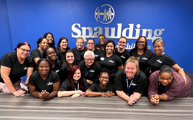 Spaulding for Children staff members smiling together for a group photo in front of the organization’s blue logo wall.